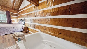 Una habitación de cabaña de madera con una bañera grande, una cama con una manta púrpura y una ventana con cortinas.