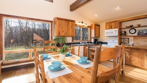 Une table de salle à manger en bois, avec de l’assiette et des chaises bleues, une cuisine avec des meubles en bois, et une vue sur les arbres à travers de grandes fenêtres.