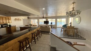 A spacious kitchen with wooden cabinets, a large island with stools, a dining area with a sofa and chairs, and a view of the outdoors through large windows. - Luxury Riverfront Home w/ Pool • Private Boat Dock • Minutes to Laughlin (Bullhead City)