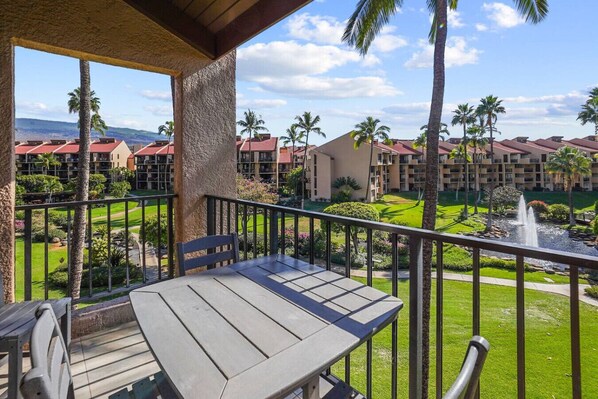 A balcony with a table and chair, overlooking a landscaped area with a fountain, palm trees, and a view of hotel buildings.