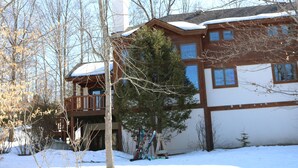 A two-story wooden cabin with a balcony, surrounded by snow and trees.