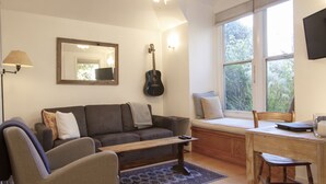 A living room with a sofa, a coffee table, a desk with a chair, a guitar, a lamp, and a window with a view of greenery.