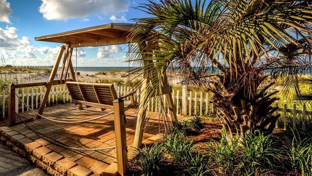 Una terraza de madera con un columpio y cerca blanca, con vistas a una playa y palmeras.