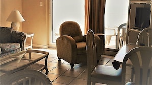 Una sala de estar con sofá, sillón y una pequeña mesa. Hay una ventana con cortinas, una lámpara y un televisor.
