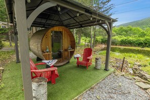 A wooden structure with a circular entrance, two red Adirondack chairs, a small table, and a bottle of water. - Sauna Belleayre Cozy Cabin (Fleischmanns)
