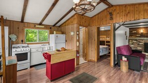 Interior rústico de una cabaña con paredes de madera, una kitchenette con estufa, refrigerador y mostrador de madera, y un rincón de estar acogedor con una silla morada.