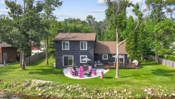 A two-story house with a dark exterior, a patio with pink chairs, and a small pond in the foreground. - Cozy cabin with 100 ft of lake shore in the heart of the brainerd lakes area! (Brainerd)