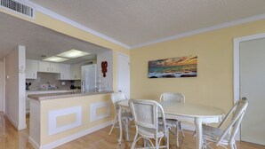 A dining area with a white table and wicker chairs, a kitchen with white cabinets, and a painting of a sunset on the wall. - Wake to the Sunrise and Steps to the Sunset! Best of Both Worlds! (Indian Rocks Beach)