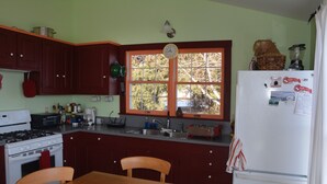A kitchen with wooden cabinets, a stove, a refrigerator, and a window with a clock. - Watch the sun rise without getting out of bed! (South Thomaston)