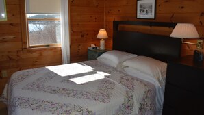 A wooden-paneled room with a bed, a nightstand, a lamp, and a window with a view of trees. - Watch the sun rise without getting out of bed! (South Thomaston)