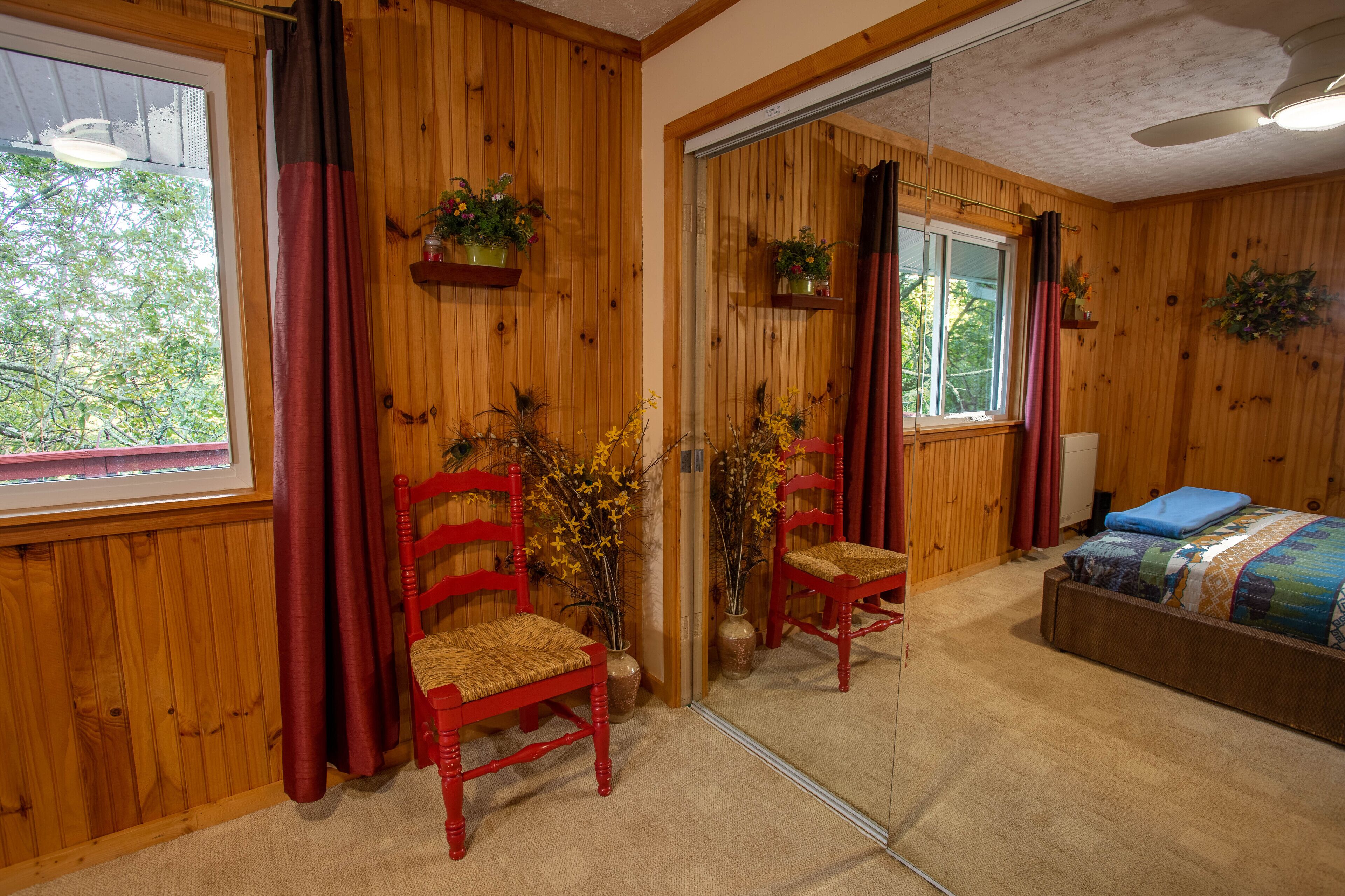 Una habitación con paneles de madera, una cama, dos sillas rojas, un espejo y plantas en macetas.