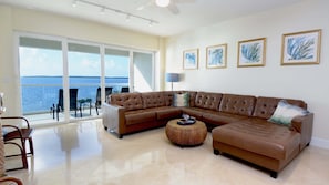 A living room with a large brown leather sofa, a round woven ottoman, and a view of the ocean through large sliding glass doors.