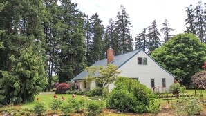 A house with a gabled roof, surrounded by a garden with various plants and flowers, and tall trees in the background.