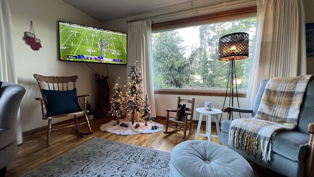 A cozy living room with a television displaying a football game, a decorated Christmas tree, and a large window with a view of trees.