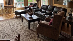 A spacious living room with a large leather sofa, a coffee table, and a patterned rug. There are multiple windows providing natural light and views of the outdoors. - Nantahala Getaway-Lakefront -mountain views. Firepit,Generator, wifi - STARLINK  (Topton)