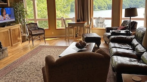 A spacious living room with a high ceiling, large windows, a television, and a cozy seating area. - Nantahala Getaway-Lakefront -mountain views. Firepit,Generator, wifi - STARLINK  (Topton)
