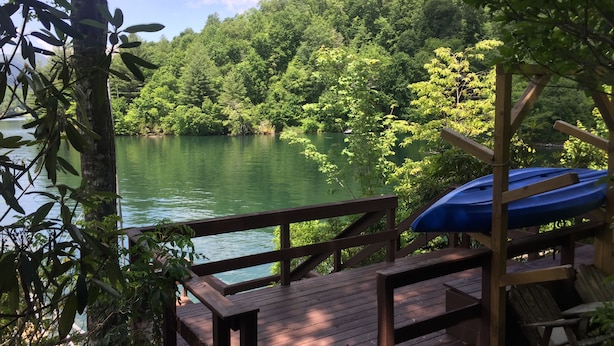 A wooden deck overlooking a lake with dense greenery and a boat on the right. - Nantahala Getaway-Lakefront -mountain views. Firepit,Generator, wifi - STARLINK  (Topton)