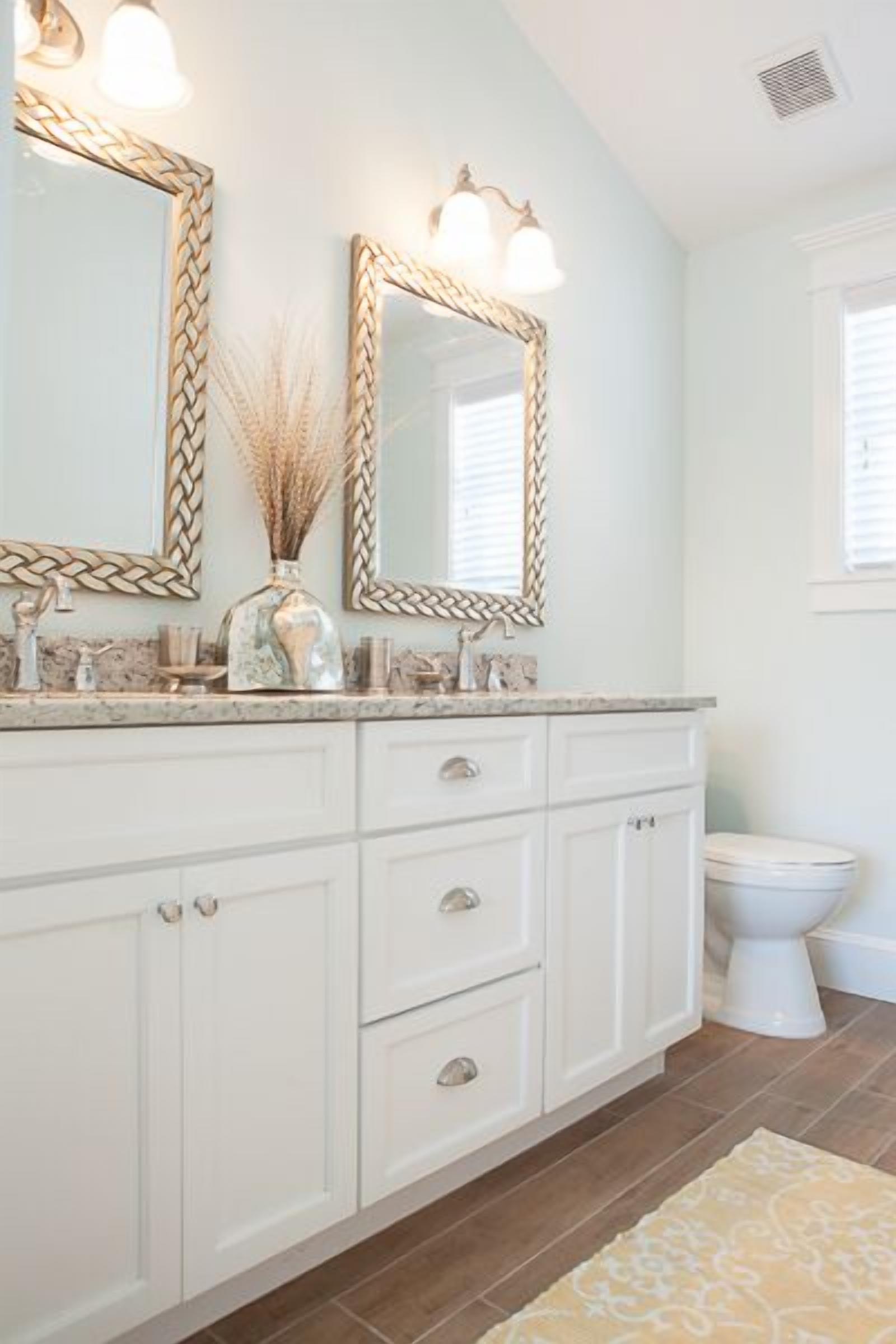 A bathroom with white cabinetry, a marble countertop, two wall-mounted mirrors with light fixtures, and a white toilet.