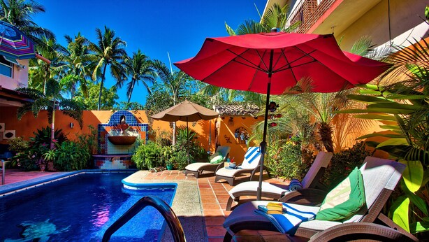 Un espace piscine aménagé avec des chaises longues et des parasols, entouré de palmiers et bordé d’une piscine carrelée.