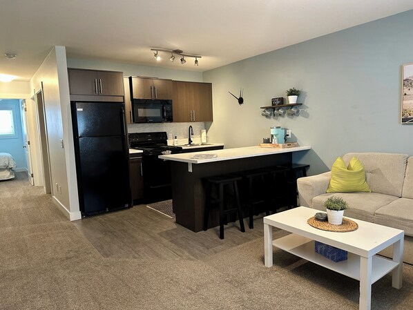 A modern kitchen with dark cabinets, a black refrigerator, and a microwave. There is a small island with stools and a coffee machine. A white table with a plant is in the living area.
