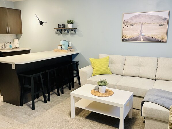 A modern living room with a kitchen area, a white sofa with a yellow pillow, a coffee table, and a road view painting on the wall.