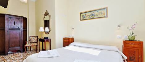 A neatly made bed with white linens, a wooden wardrobe, a chair with a floral cushion, a wall-mounted mirror, and a framed picture.