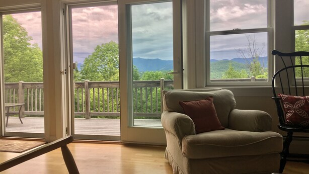 Ein Zimmer mit einem beigen Sofa, einem roten Kissen und Blick auf einen Balkon mit Holzgeländer.