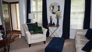 A living room with a white armchair, a blue ottoman, and a beige sofa. There is a small table with a lamp and a mirror on the wall. - Modern 4BR Retreat — Minutes from Mercedes-Benz Stadium - World Cup Ready (Atlanta)