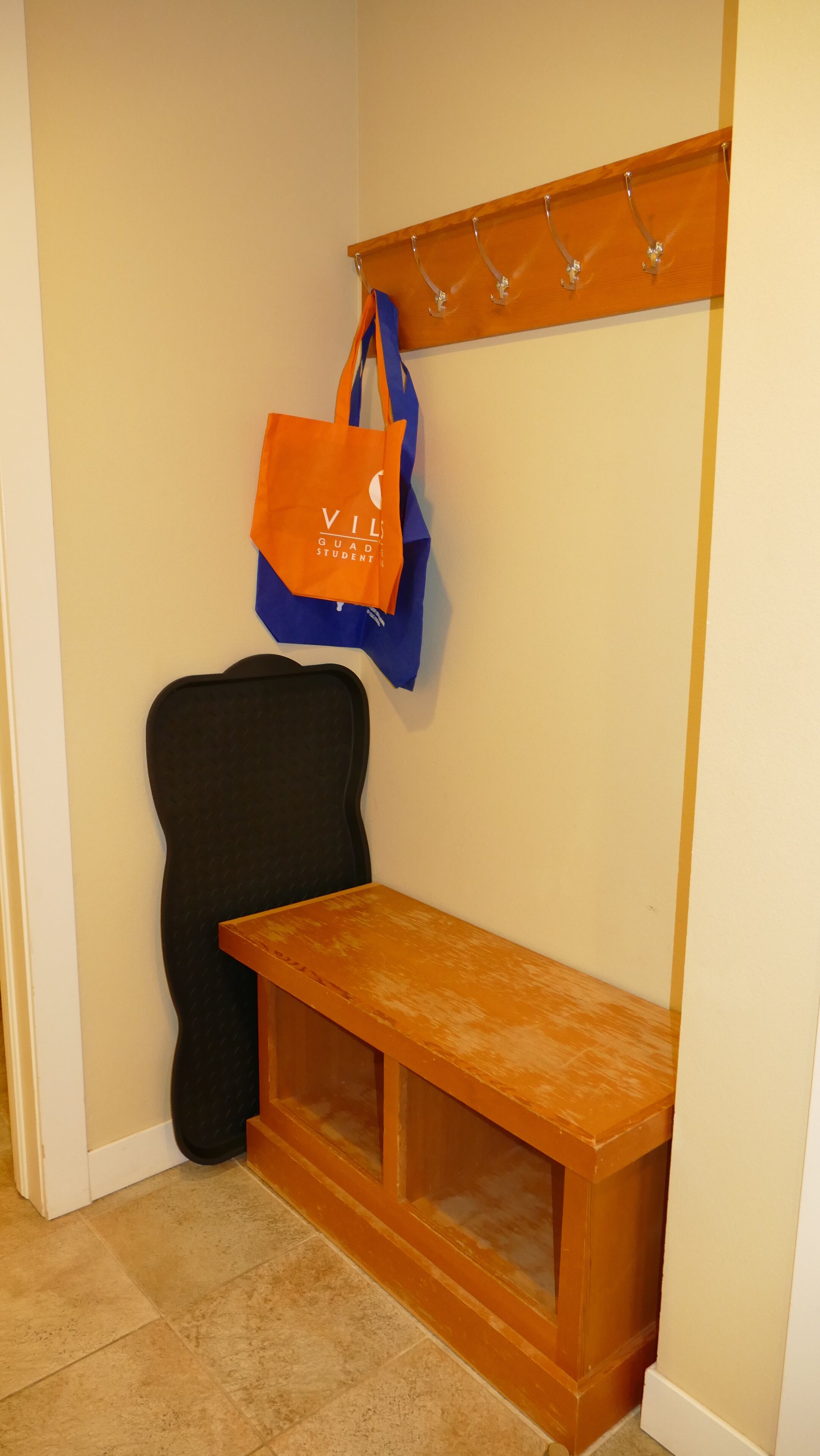 Eine Sitzecke mit einer hölzernen Bank, einem schwarzen Stuhl und einer orangefarbenen Tasche, die an einem Metallgestell hängt.