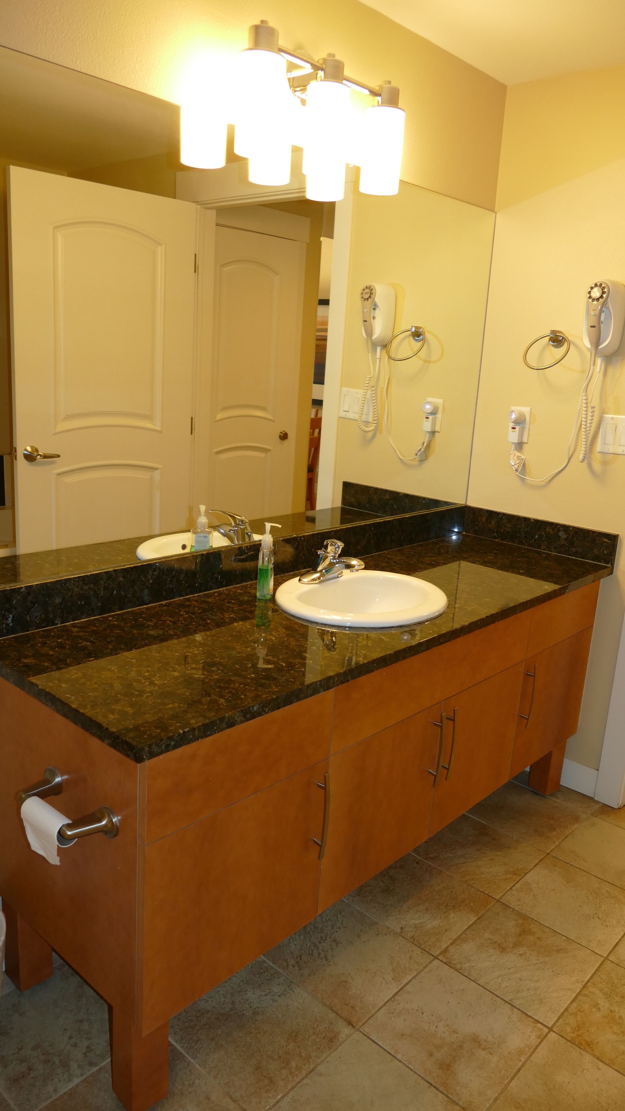 A bathroom with a double sink, vanity, and a mirror. There are two hair dryers mounted on the wall.