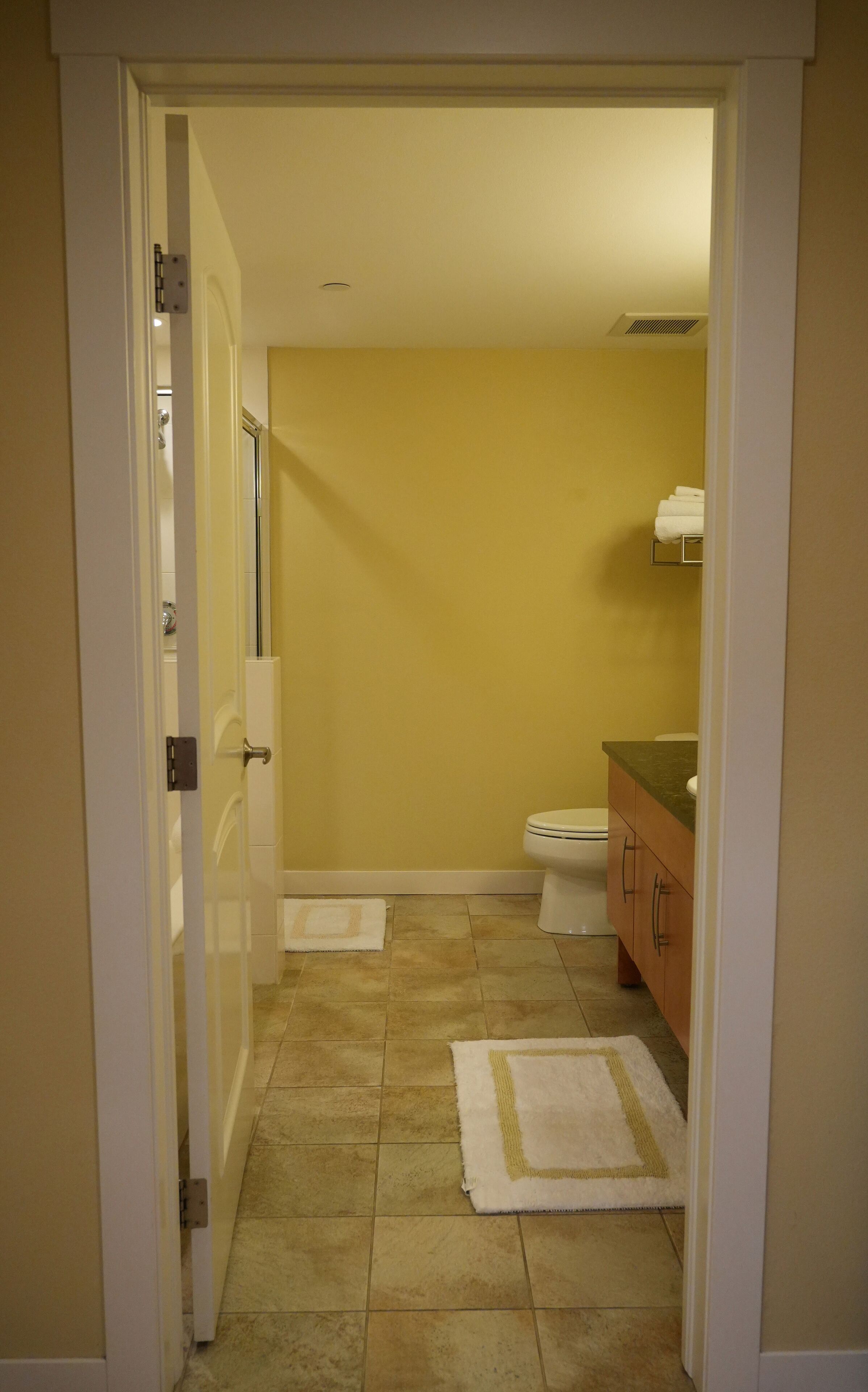 Un couloir étroit mène à une salle de bain aux murs jaunes, équipée de toilettes et d’un lavabo.