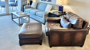 A living room with a light blue sofa, a brown leather couch, a coffee table, and a ceiling fan. There is a painting on the wall and a lamp on a side table. - Ocean Front Amelia Island, Beachfront Pool, 20% Off Sunglasses at Eye Envy store (Fernandina Beach)