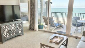 A living room with a TV, a coffee table, and a sofa. There is a balcony with a view of the ocean. - Ocean Front Amelia Island, Beachfront Pool, 20% Off Sunglasses at Eye Envy store (Fernandina Beach)