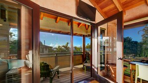 A modern room with a sliding glass door leading to an outdoor deck, a flat-screen TV mounted on the wall, and a small table with chairs. - Ocean view Steps to Hanalei Beach TVNC#5087 (Hanalei)