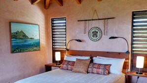 A bedroom with a large bed, wooden ceiling beams, a ceiling fan, a painting of a landscape, two bedside lamps, and a decorative wall hanging. - Ocean view Steps to Hanalei Beach TVNC#5087 (Hanalei)