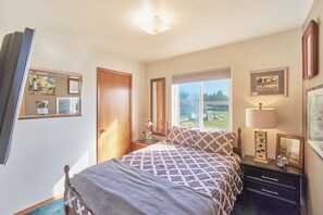 Een slaapkamer met een bed, nachtkastje, lamp, dressoir en een raam met uitzicht op de omgeving.