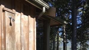 Eine Holzterrasse führt zu einem überdachten Bereich mit einer Metalttür, umgeben von Bäumen und einem See.