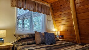 A bedroom with a large bed, a window with blinds, a lamp on a bedside table, and wooden walls.