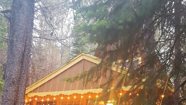 A wooden deck with a dining table and chairs, surrounded by trees and a building with string lights.
