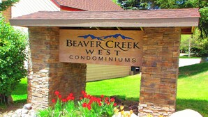 A sign for Beaver Creek West Condominiums with a stone structure and a grassy area with red flowers. - Remodeled Condo w/ FREE WiFi, Parking, Heated Pool, Hot Tubs, Skier Shuttles (Avon)