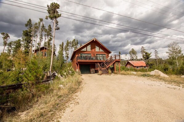A two-story log cabin with a balcony, surrounded by trees and a dirt driveway.