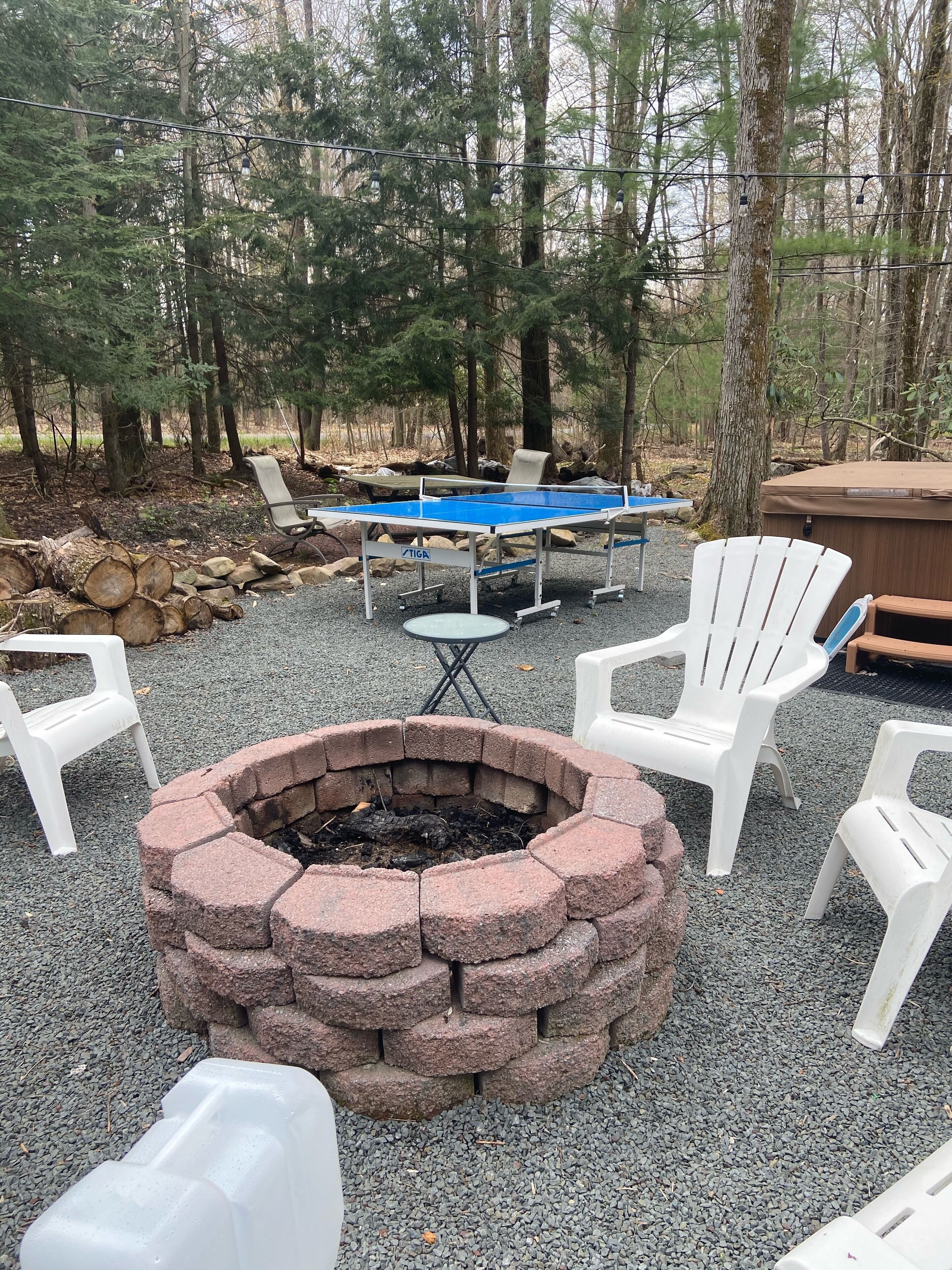 A fire pit surrounded by white chairs and a table with chairs, set in a forested area with trees and a wooden bench in the background.