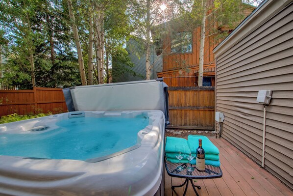 A hot tub with a wooden deck, a table with a bottle and glasses, and a wooden fence in the background.