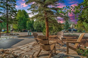 An outdoor seating area with Adirondack chairs, a stone pathway, and a backdrop of trees and a sunset sky.