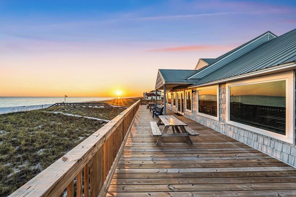 A wooden deck with a view of the ocean at sunset, a covered outdoor seating area, and a building with large windows.