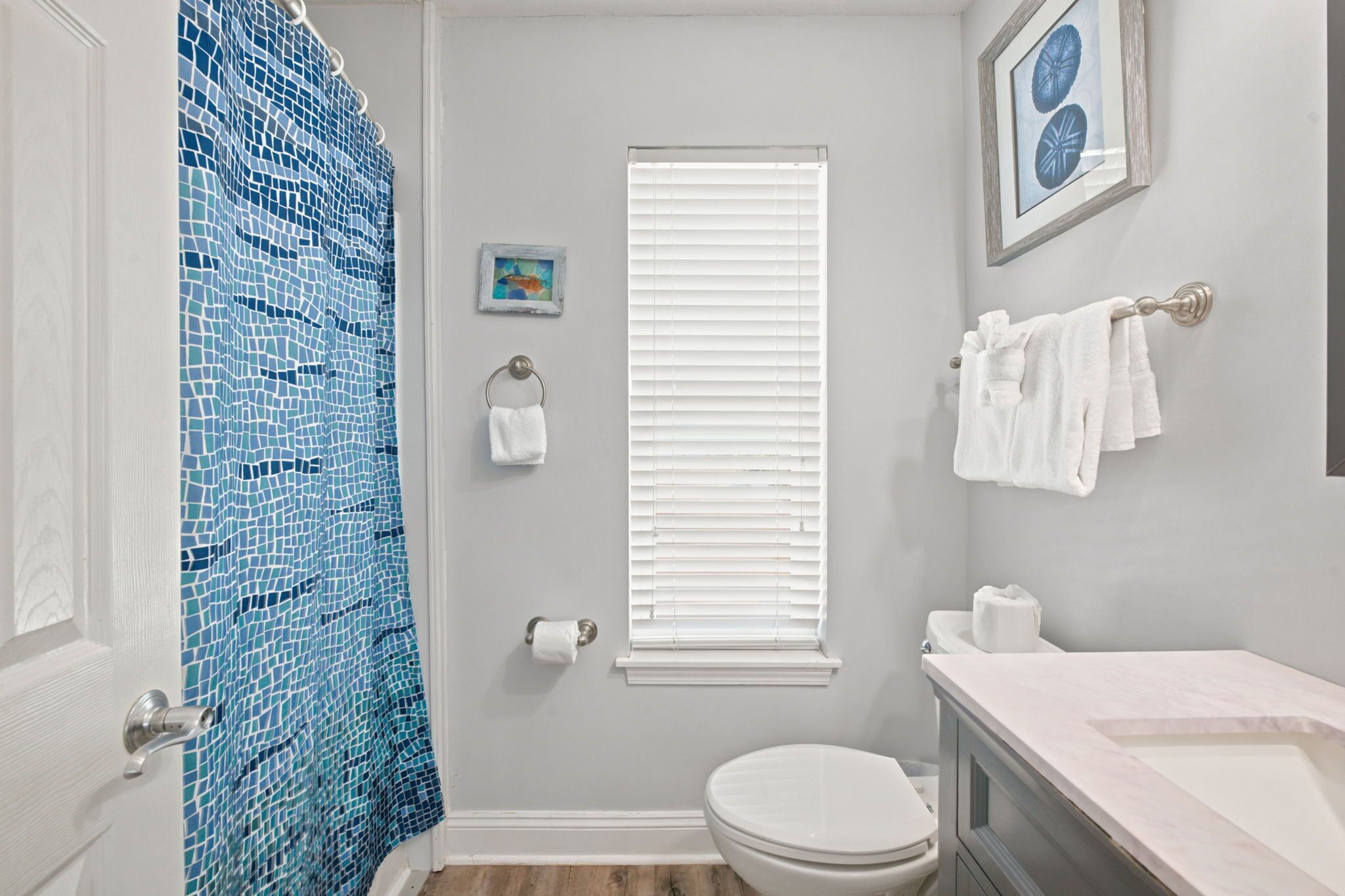 Un bagno con il water, un lavandino, una tenda da doccia, una finestra con persiane, un portasciugamani e un quadro appeso al muro.
