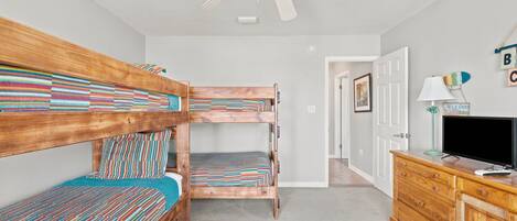 A room with bunk beds, a ceiling fan, a television on a wooden dresser, and a door leading to another room.