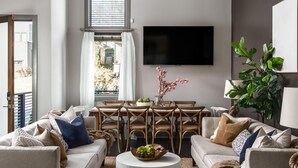 Een moderne woonkamer met een eethoek, een flatscreen televisie aan de muur en een groot raam met gordijnen.