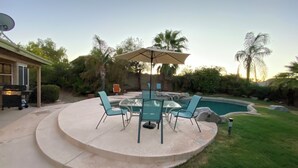 A circular patio with a glass table and chairs, a barbecue grill, and a swimming pool in the background.