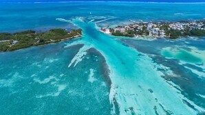 An aerial view of a coastal area with clear turquoise waters, a small island with buildings, and a narrow waterway connecting them. - Oceanfront home; Rooftop deck; Fabulous views; Upper level apartment (Caye Caulker)